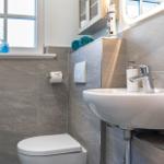 Badezimmer mit Toilette, Waschbecken und Fenster. Graue Fliesen und weißer Spiegel.