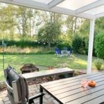 Terrasse mit Tisch, Bank und Grill. Im Hintergrund Garten mit Sitzgruppe und Bäumen.