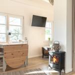 Ein Raum mit Esstisch, Fernseher und Kaminofen. Durch Fenster fällt Tageslicht.