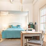 Schlafzimmer mit blauer Bettdecke, Schreibtisch und großen Fenstern.