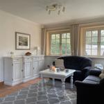 Wohnzimmer mit zwei dunklen Sofas, weißem Tisch und Fenstern mit Vorhängen.