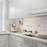 Küche mit weißen Schränken, Arbeitsfläche und Herd. Fenster, Spüle und Küchenutensilien sind sichtbar.