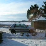 A silver car is parked on a snow-covered property beside trees and a fence.