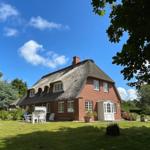Rotes Backsteinhaus mit Strohdach und weißem Eingang. Grünflächen, Bäume und Gartenmöbel.