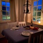 Esstisch mit Tischdecke, Gläsern, Wein und Käse vor Fenstern mit Blick auf Garten.