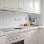 Küche mit weißen Schränken, Herd und Spüle. Auf der Arbeitsfläche liegen Obst, Gemüse und ein Buch.