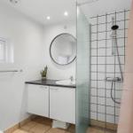 Bathroom with shower, sink and toilet. White walls, round mirror and brown tiles.