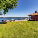 Rotes Bootshaus am See mit Terrasse und Liegeplatz. Grasfläche und Baum im Vordergrund.