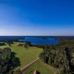 Aerial view of a lake surrounded by forests and green fields with a house nearby.