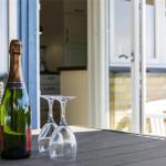 Eine Flasche Sekt und zwei Gläser stehen auf einer Terrastisch. Im Hintergrund ist eine Küche und ein Balkon sichtbar.
