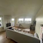 Wohnraum mit Sofa, Kamin und Fenstern zum See. Holzboden und Wandverkleidung.