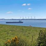 Green field with view of sea, shipyard, and wind turbines in the background.
