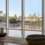 Durch große Fenster blickt man auf eine natürliche Landschaft mit Bäumen und Sträuchern.