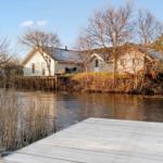 Haus am Wasser mit Holzsteg und Terrasse. Bäume und Schilf umgeben die ruhige Umgebung.