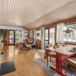 A room with dining area, living area, and wood ceiling. It has large windows and a fireplace.