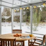 A wooden table with chairs, oranges, and a cup under a glass roof with string lights.