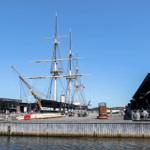 Historic sailing ship at the harbor with modern buildings and visitors.