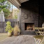Outdoor area with stone fireplace, table, chairs, and plants. Background: wooden fence and trees.