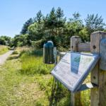 Sign and trash bin along a wooded path with grassy hills and trees.
