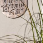 Rundes Holzschild mit Schriftzug und Hausmotiven an Wand, Gräser im Vordergrund.