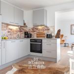 Küche mit Backofen, Arbeitsplatte und Backsteinwand. Blick in Wohnbereich mit Sessel und Tisch.