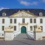 Eingangsbereich mit Brücke und Löwenstatuen vor einem weißen Gebäude mit dunklem Dach.