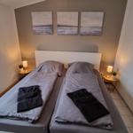 Zwei Betten mit grauer Bettwäsche und schwarzen Handtüchern. An der Wand hängen drei Bilder.