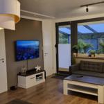 Wohnzimmer mit Sofa, Couchtisch und Fernseher. Große Fenster mit Blick auf Terrasse.