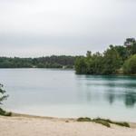 Ein See mit sandigem Ufer und umgebenden Bäumen unter einem bewölkten Himmel.