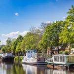 Schiffe liegen am Kanal, umgeben von Bäumen und Häusern unter blauem Himmel.