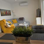 Wohnzimmer mit Sofa, gelber Sessel und TV. Fenster mit Jalousien.