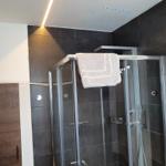 Duschkabine mit Glaswand und Duschbrause. Dunkle Fliesen und Deckenbeleuchtung.
