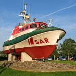 SAR-Schiff als Skulptur auf grünem Rasen mit Steinen und Bäumen im Hintergrund.