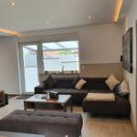 Wohnzimmer mit Sofa, Couchtisch und TV-Wand. Fenster mit Blick auf Garten.