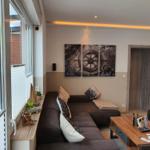 Wohnzimmer mit Sofa, Couchtisch und Wandbildern. Große Fenster mit Blick auf Außenbereich.