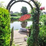 Durch einen Bogen führt ein Weg zu einer Terrasse mit Liege und Sonnenschirm am Wasser.