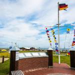 Marineflaggenmast mit deutscher Flagge und Informationsplaketten vor Hafen.