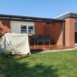 Terrasse mit Tisch, Stühlen und Grill unter Pavillon. Rasen und Blumen im Vordergrund.