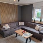 Ein Wohnzimmer mit grauem Sofa, Holztisch und Fenster mit Pflanzen.