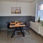 Esszimmer mit Holztisch, vier Stühlen und Wandbild. Fenster mit Pflanzen.