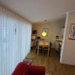 Wohnung mit Essbereich, Küche und Kaminofen. Durchgehende Holzdielen und weiße Vorhänge.