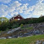 Rotes Haus auf grüner Anhöhe mit Steinhang und blauem Himmel.