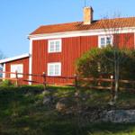 Rotes Holzhaus mit weißem Rahmen und Ziegeldach. Umgeben von Gras, Bäumen und einem Holzzaun.
