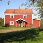 Rotes Holzhaus mit grüner Tür und rotem Dach unter Bäumen.