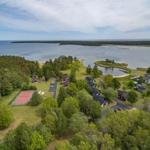 Luftbild einer Ferienanlage am Wasser mit Häusern, Bäumen und einem Tennisplatz.