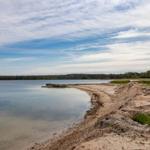 Strandufer mit Sand, Bäumen und ruhigem Wasser im Hintergrund.