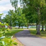Weg durch grüne Bäume zum See. Ferienhaus links. Picknicktische im Gras.