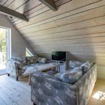 Wohnzimmer mit Holzwänden, Sofa und Fernseher. Türe führt auf Balkon mit Blick in Garten.