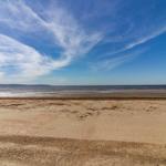 Strand mit Sand und flachem Wasser unter blauem Himmel mit weißen Wolken.
