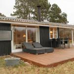 Holzterrasse mit Liegen und Tisch. Haus mit großen Fenstern und Schornstein.